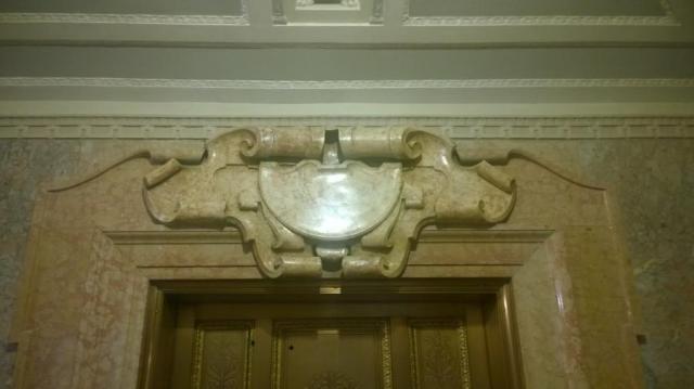 Cast scagliola cartouche over elevator doors, matching the adjacent molded marble surrounds.